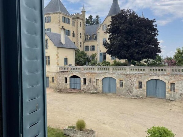 A courtyard with a large building featuring a tower and a tree. There are multiple doors and a stone wall.