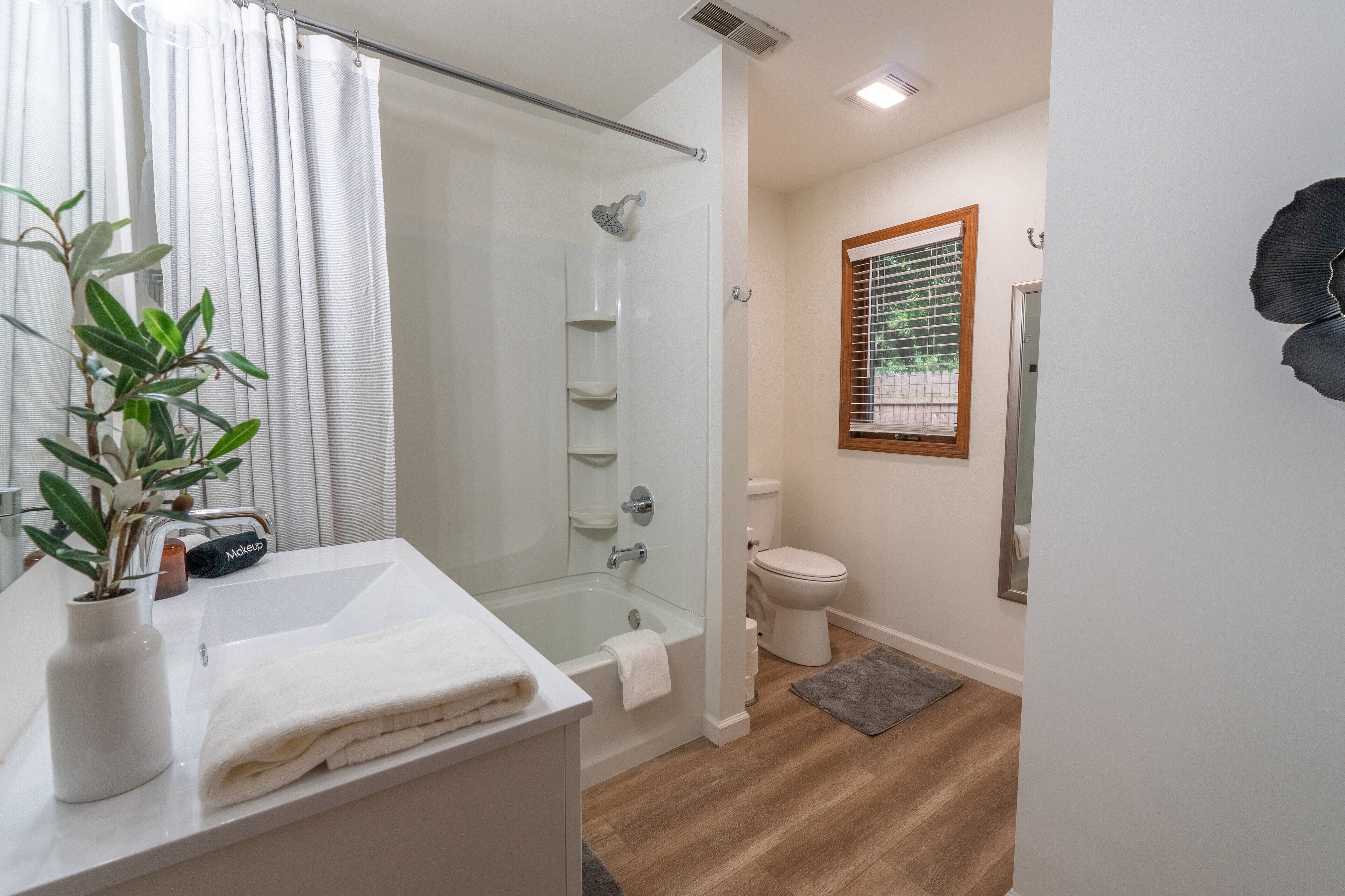 A modern bathroom with a glass shower enclosure, a white sink with a vase of green plants, and a toilet with a wooden-framed window.