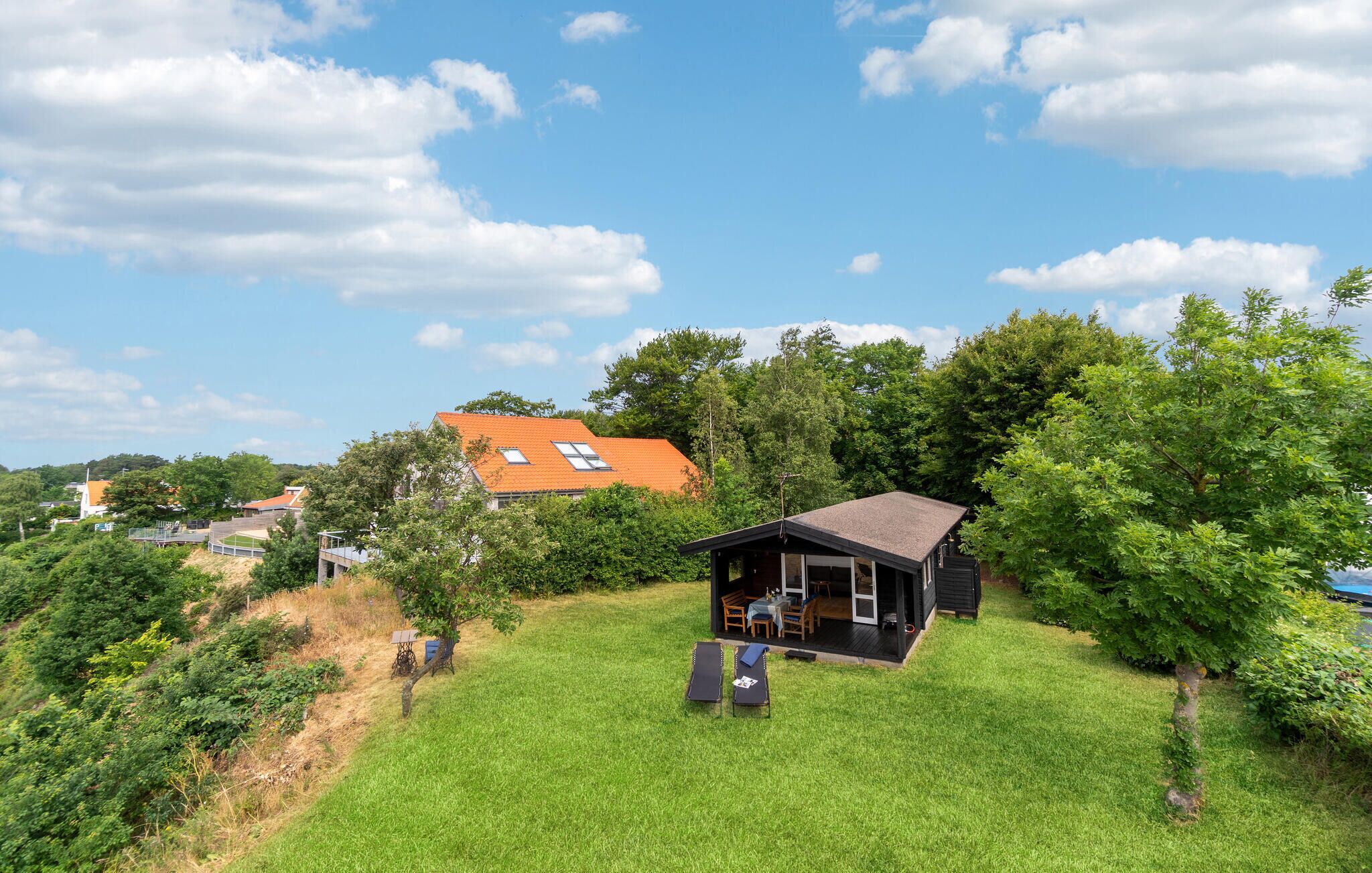 A small house with a dark roof and a porch, surrounded by lush greenery and a clear blue sky.