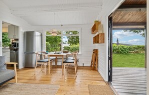 A dining area with a wooden table and chairs, a kitchen in the background, and a view of greenery outside.