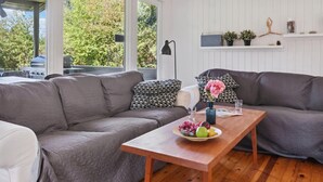 A cozy living room with a grey sofa, a wooden coffee table with a bowl of fruit and flowers, and a white wall with a shelf holding plants and decorative items. - 2 bedroom gorgeous home in Askeby (Askeby)