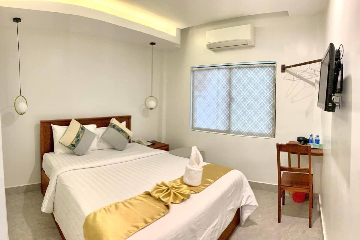 A hotel room with a bed, a desk, a chair, a TV, and an air conditioning unit.