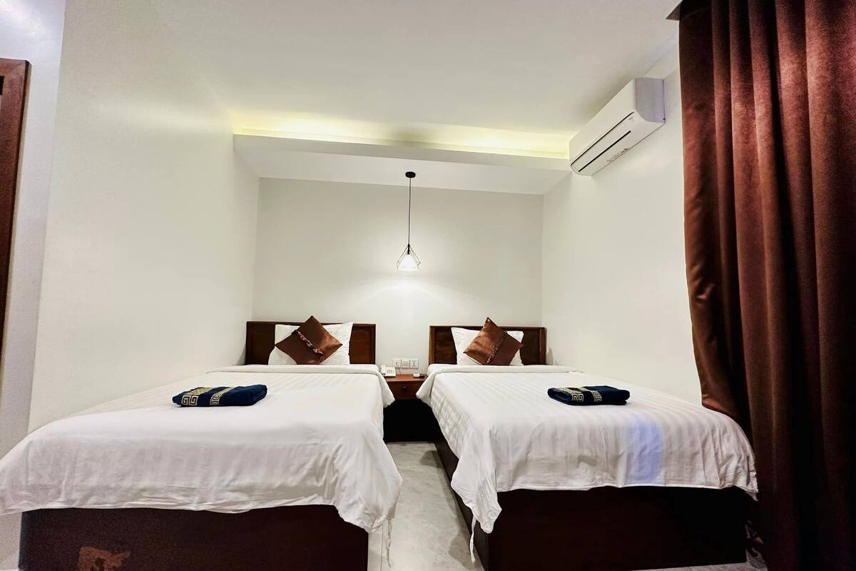 A hotel room with two single beds, a small table, a hanging light fixture, and an air conditioning unit.