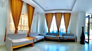 A modern living room with a sectional sofa, a standing table, and a large window with curtains. - Laluna Guesthouse (Siem Reap)
