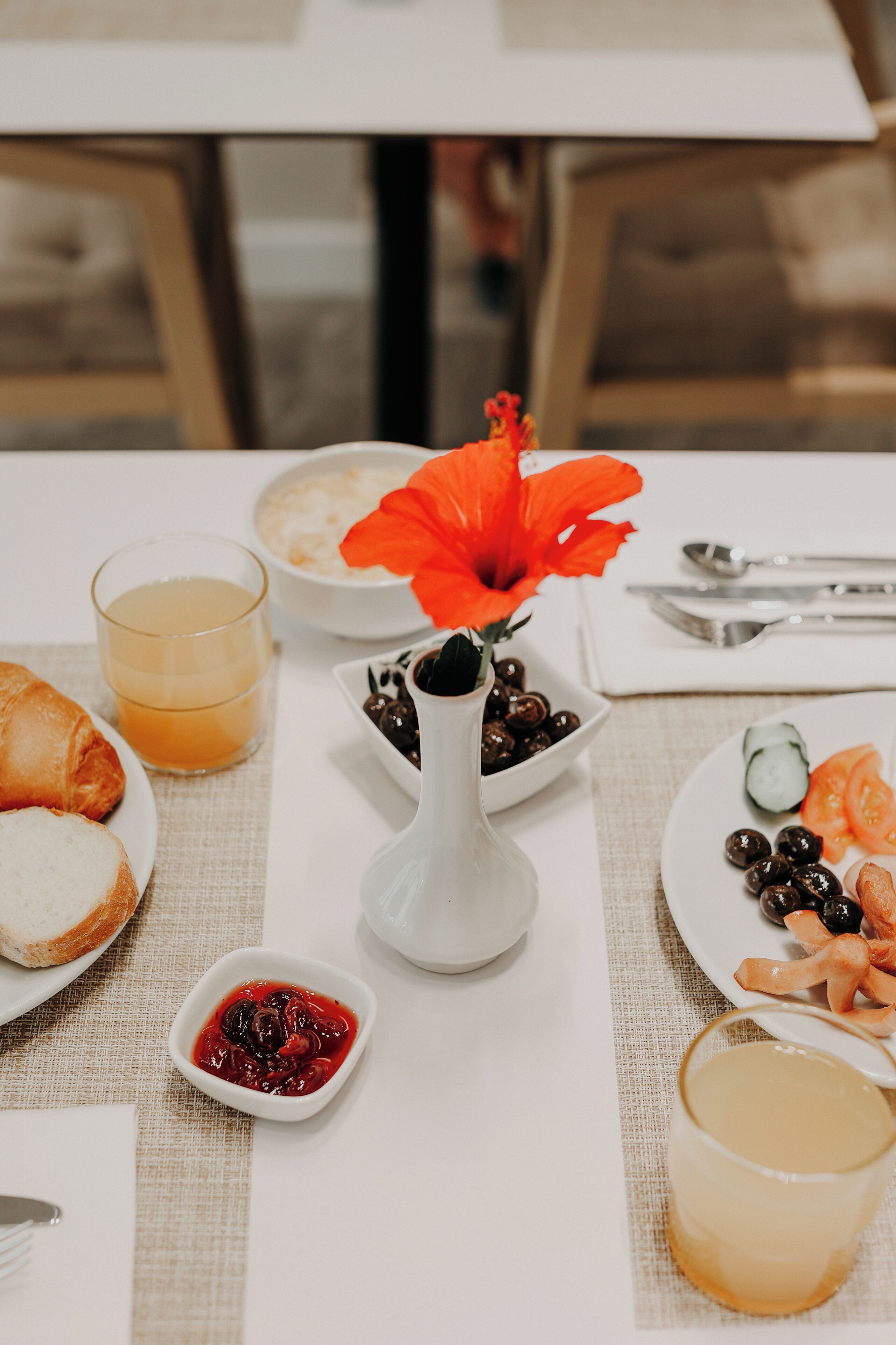 Una mesa de desayuno con cruasanes, un cuenco de aceitunas, un vaso de zumo de naranja, un plato con tomates y pepinos, y un florero con una flor roja.