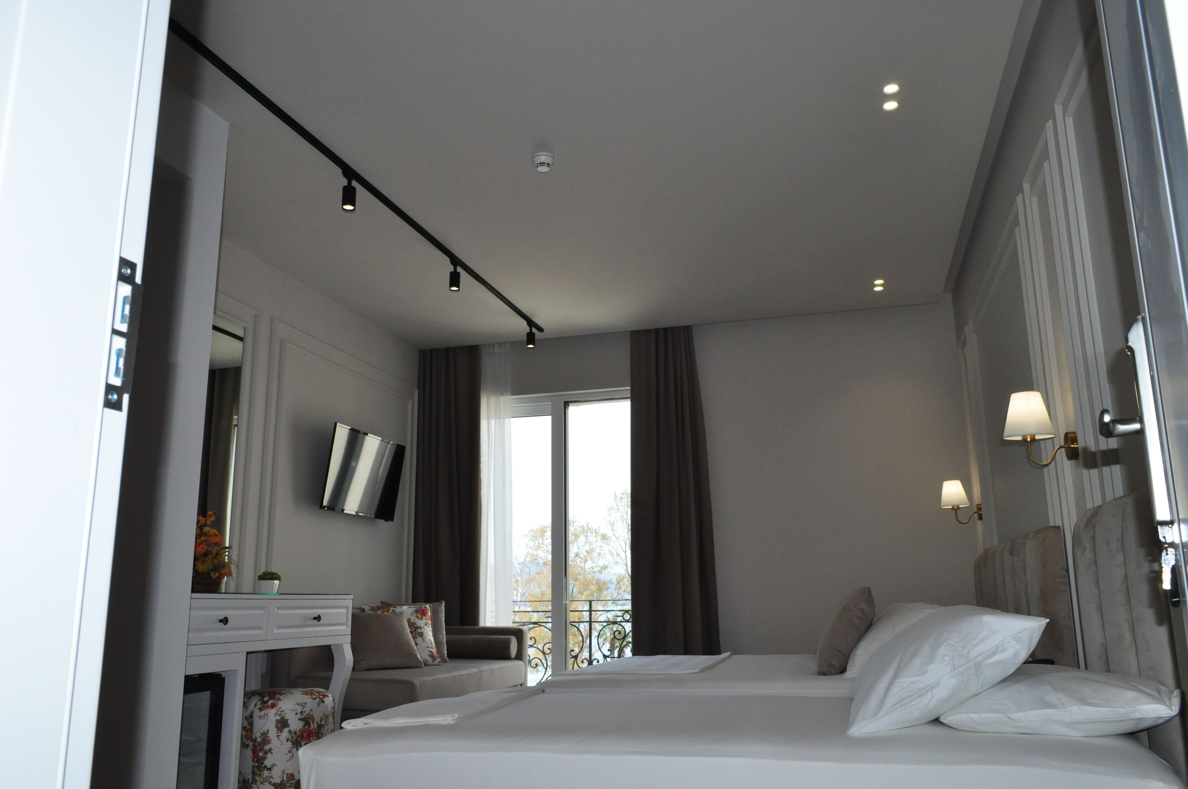 A hotel room with a bed, a desk with a flat-screen TV, a window with curtains, and a balcony view.