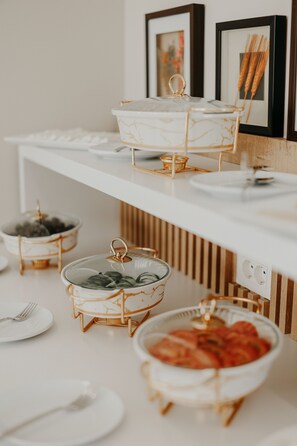 A buffet setup with various food containers on a white table, framed pictures on the wall, and a power outlet.