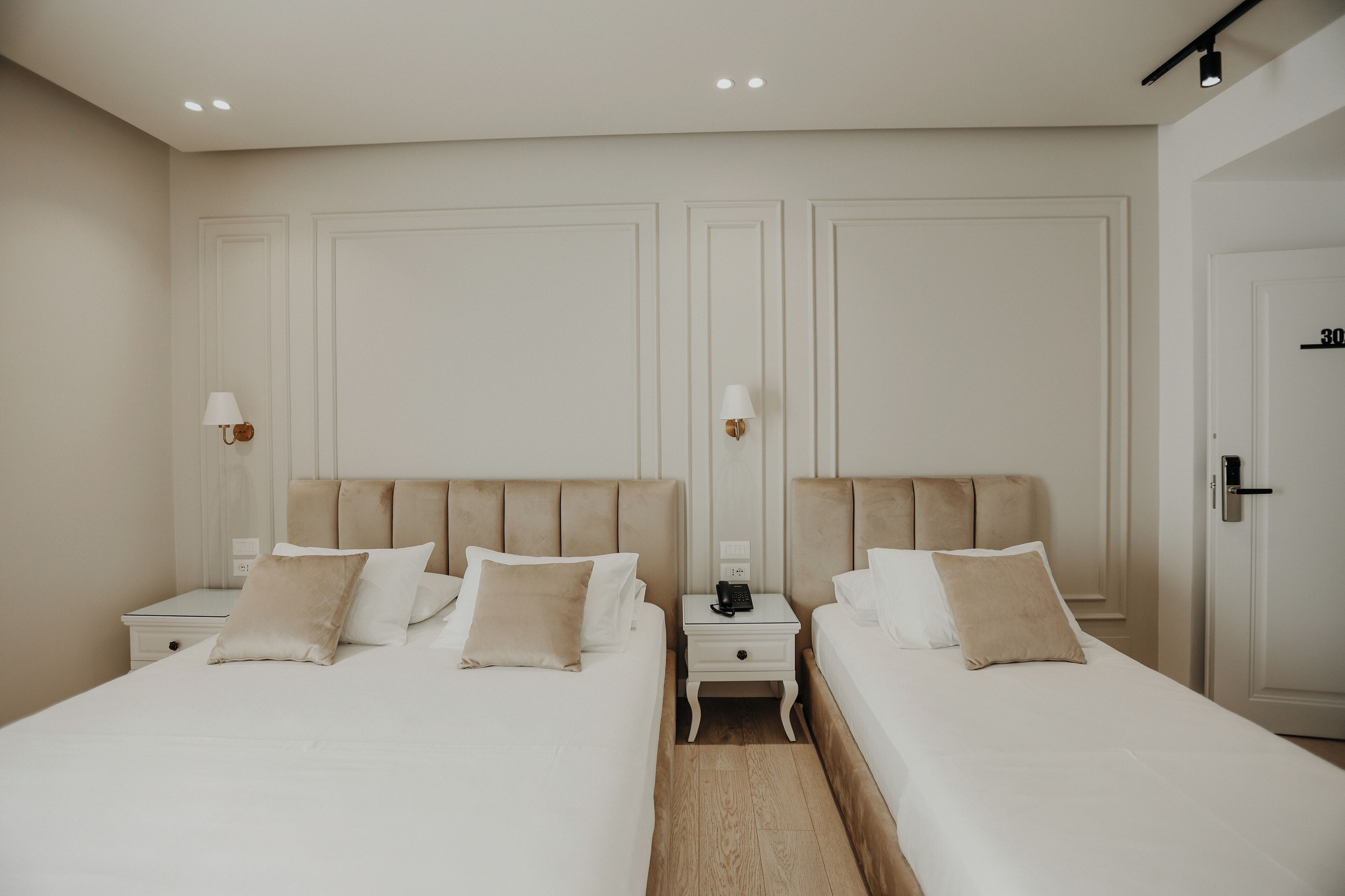 A hotel room with two beds, a nightstand with a lamp, and a door with a number.