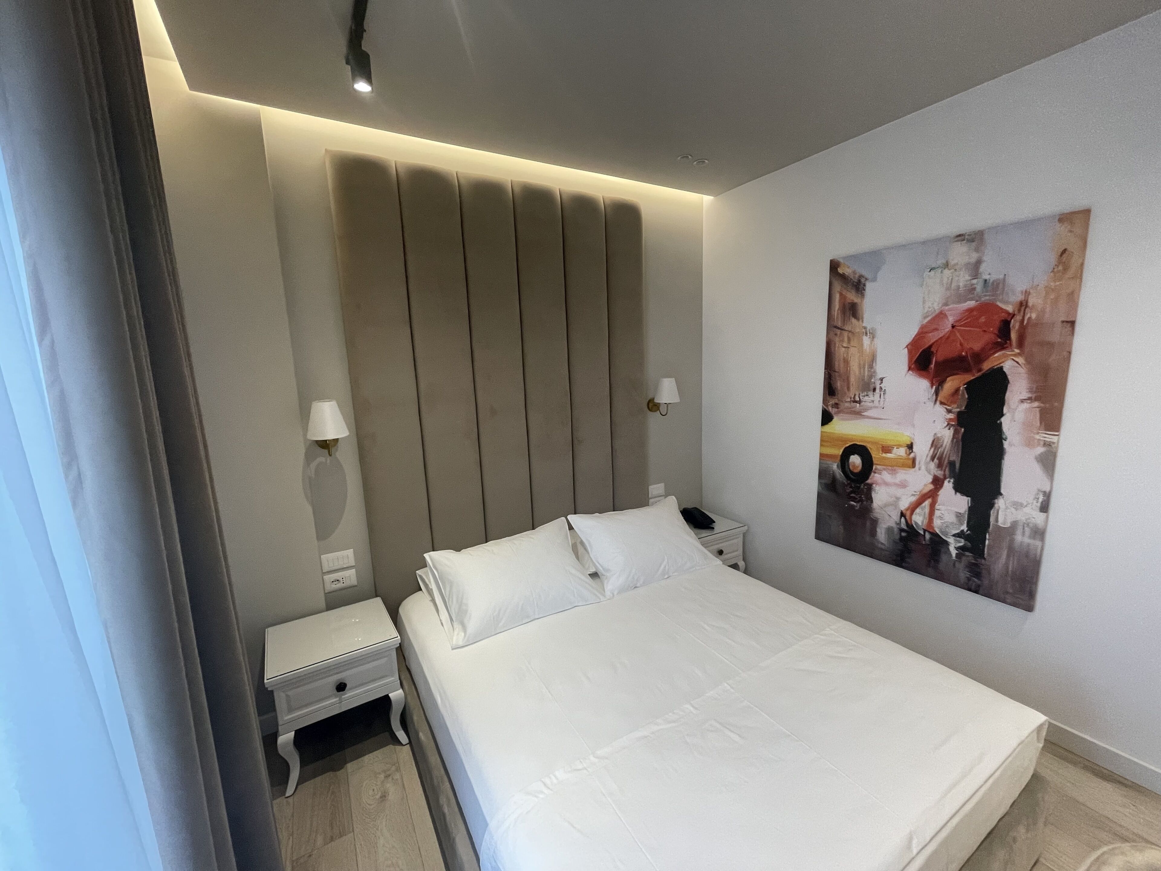 Een moderne hotelkamer met een groot bed, nachtkastjes, een wandlamp en een schilderij van een regenachtig straatbeeld.