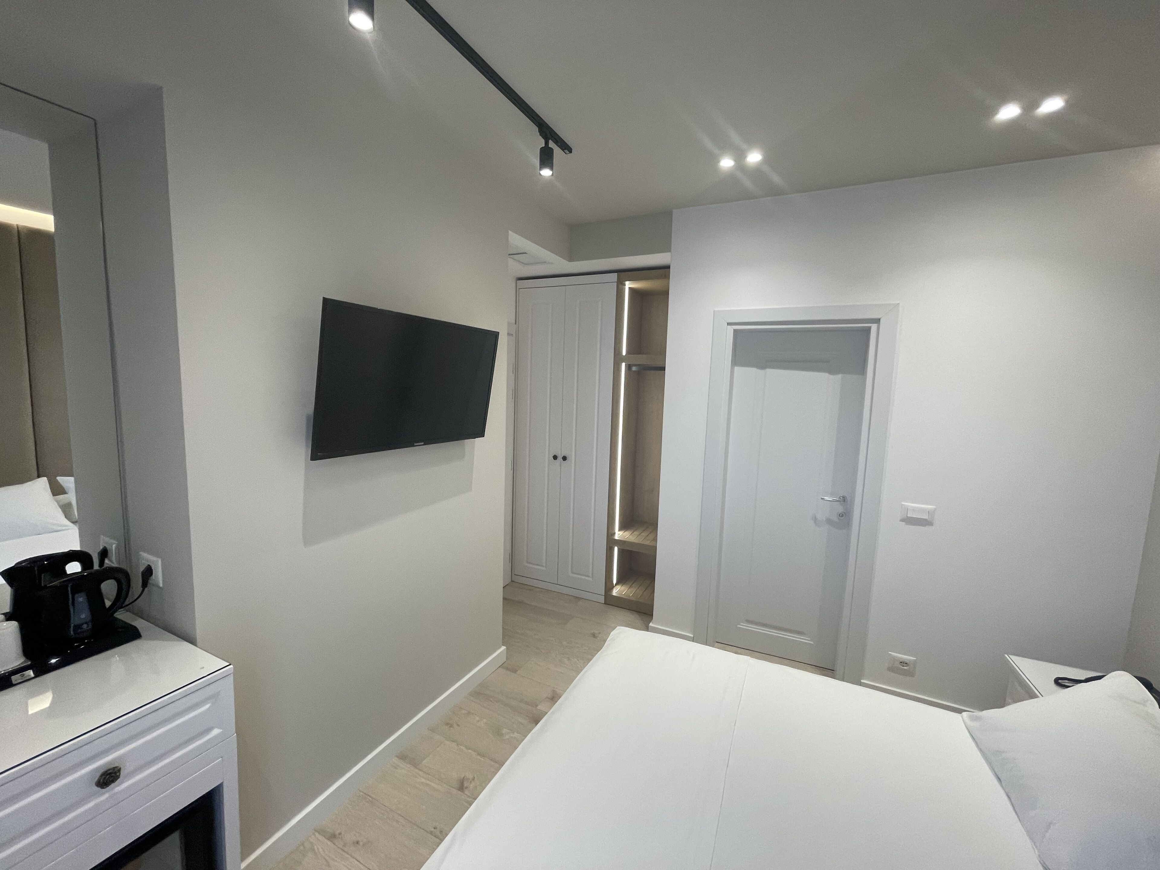 A modern hotel room with a bed, a TV mounted on the wall, a coffee maker on a bedside table, and a wardrobe with a mirror.