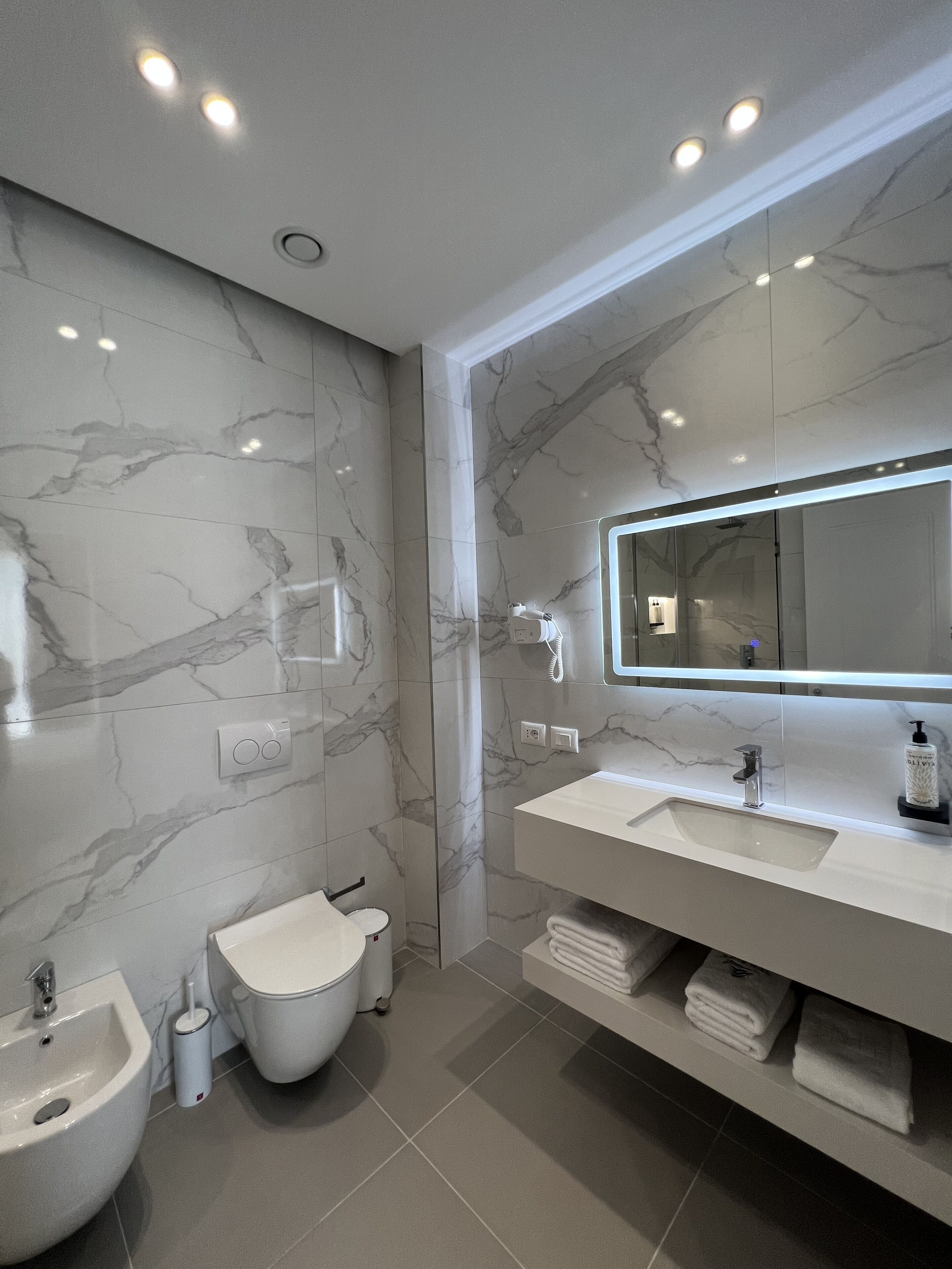 A modern bathroom with marble walls, a large mirror, a bidet, a sink, and a towel rack.
