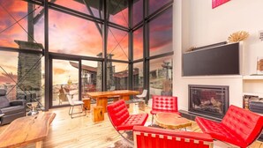 A spacious living area with large windows, a wooden table, red chairs, and a fireplace.