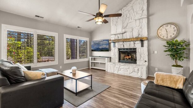 A modern living room with a stone fireplace, large windows, and a ceiling fan.