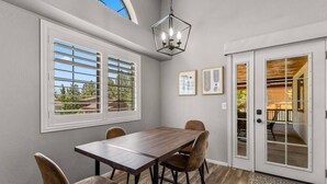 A dining area with a wooden table and chairs, large windows with blinds, a chandelier, and a sliding glass door leading to an outdoor patio.