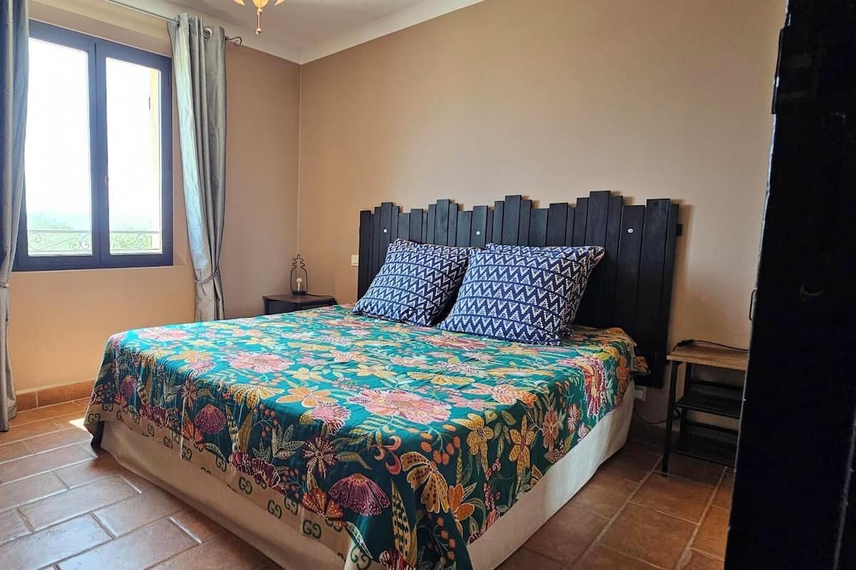 Un dormitorio con cama, mesita de noche, silla, ventana con cortinas y un cuadro en la pared.