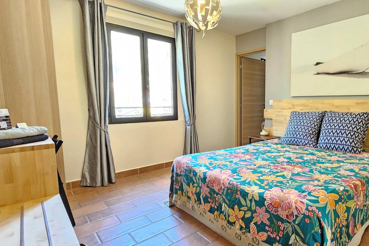 Un dormitorio con una colcha estampada, cabecera de madera, mesita de noche y ventana con cortinas.
