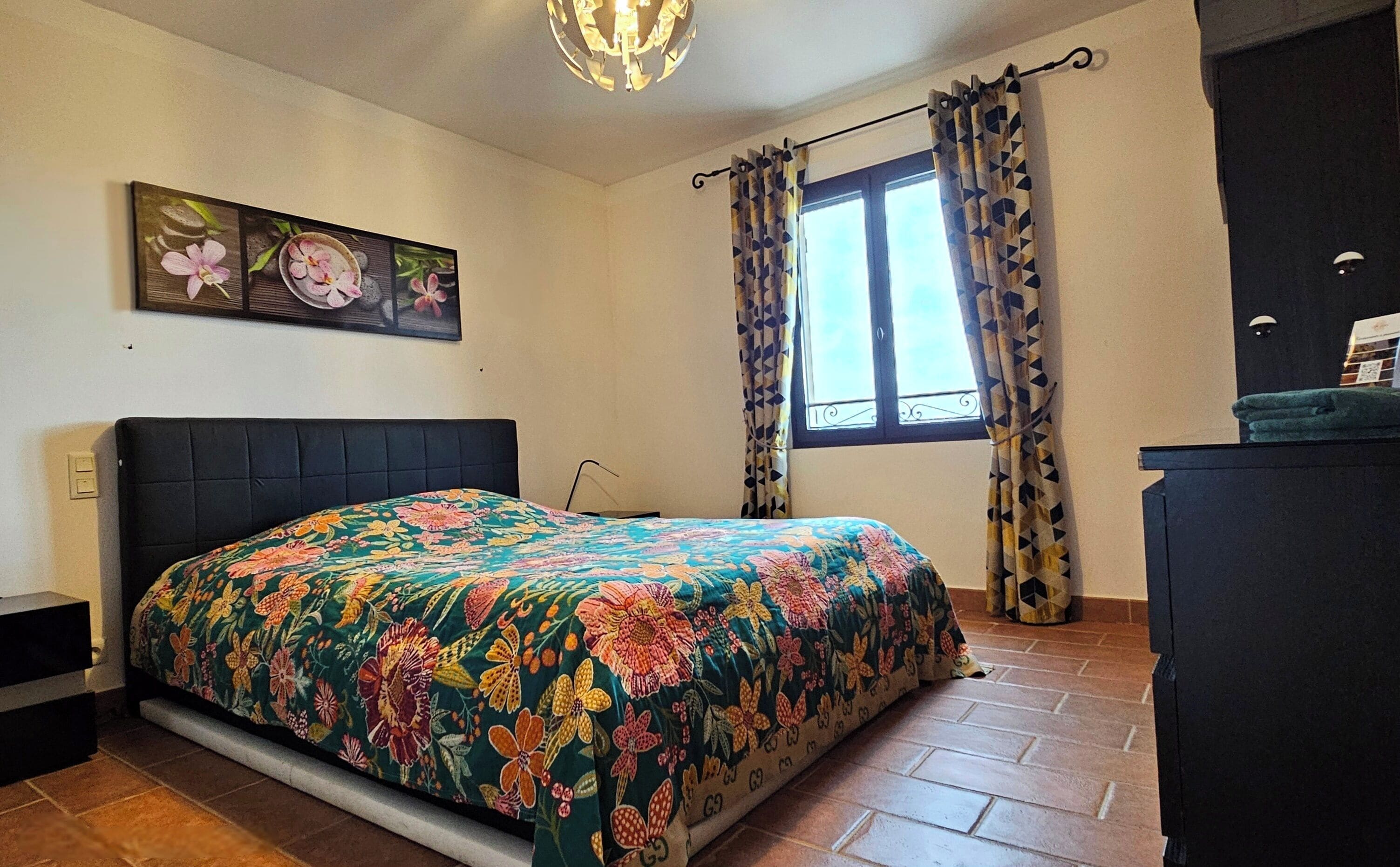 Un dormitorio con colcha estampada, una ventana con cortinas decoradas y un cuadro en la pared.