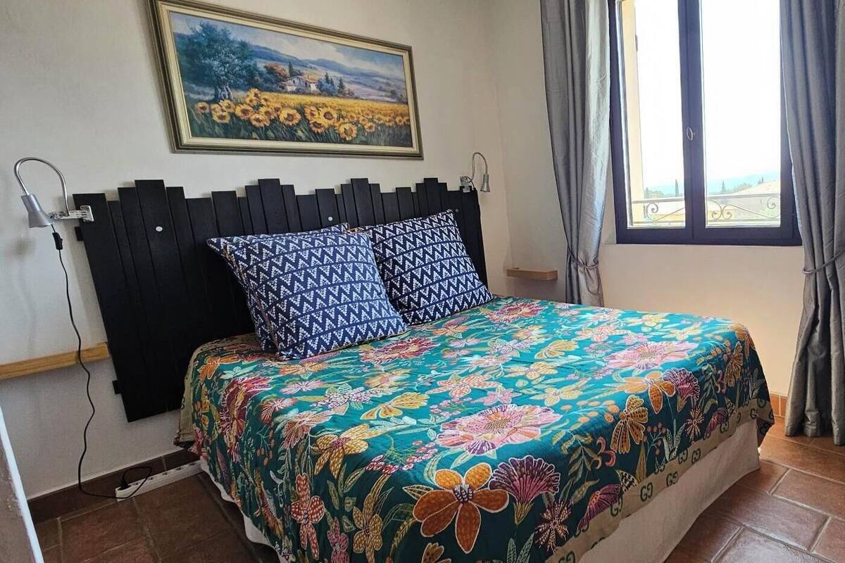 Un dormitorio con cama, un cuadro, una ventana con cortinas y una mesita con un cartel.