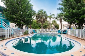 Piscina com cerca branca, rodeada por árvores e um edifício com esquema de cores azul e branco.