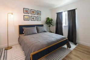 A bedroom with a bed, a floor lamp, a window with curtains, and framed pictures on the wall.