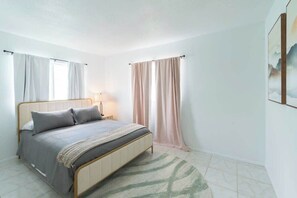 A bedroom with a bed, curtains, a lamp, and a painting on the wall. - Fillmore Unit 3 | 2BR/1BA (Cape Canaveral)