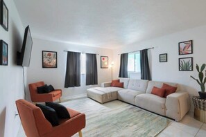A modern living room with a sectional sofa, a single armchair, and a flat-screen TV mounted on the wall. - Fillmore Unit 3 | 2BR/1BA (Cape Canaveral)