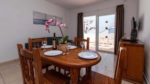 A dining area with a wooden table set for four, a vase of pink flowers, and a bottle of wine. - Las Floritas 22 - One Bed ground floor with pool (Playa de la Américas)