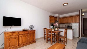 A compact living space with a wooden dining table, a TV mounted on the wall, and a kitchenette with wooden cabinets. - Las Floritas 22 - One Bed ground floor with pool (Playa de la Américas)