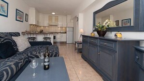 A modern living room with a sofa, a coffee table with a bottle and glasses, a kitchen in the background, and a large mirror above the coffee table. - Santa Maria 67 - Two Bed (Costa Adeje)