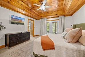 A bedroom with a wooden ceiling, a bed with a white quilt, a dark dresser, a television on the wall, and a window with curtains.