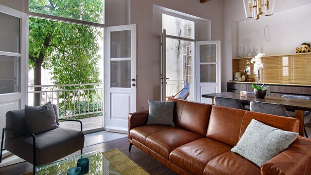 A spacious living room with a brown leather sofa, a glass coffee table, and a grey armchair. Large windows offer a view of a balcony with greenery.