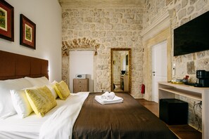 A hotel room with a stone wall, a bed with white and yellow pillows, a flat-screen TV, and a wooden nightstand. - Central W Apartment and Rooms by DuHomes (Dubrovnik)