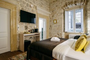 A bedroom with stone walls, a flat-screen TV, a desk with a coffee maker, and a window with a view. - Central W Apartment and Rooms by DuHomes (Dubrovnik)