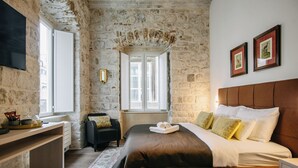 A bedroom with a stone wall, a bed with white and brown bedding, a desk with a lamp, and a window with a view.