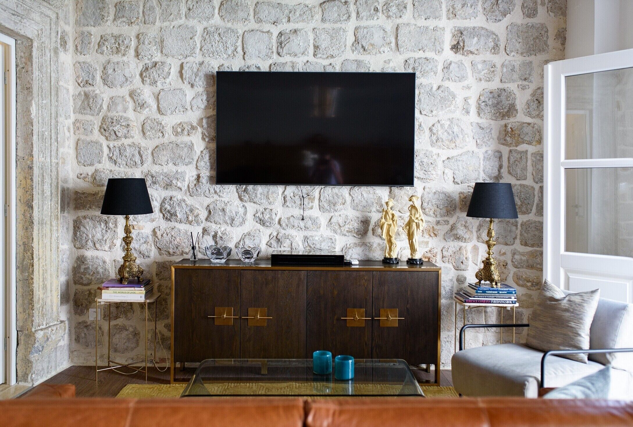 A living room with a stone wall, a flat-screen TV mounted above a wooden cabinet, two floor lamps, a glass coffee table, and a leather sofa.
