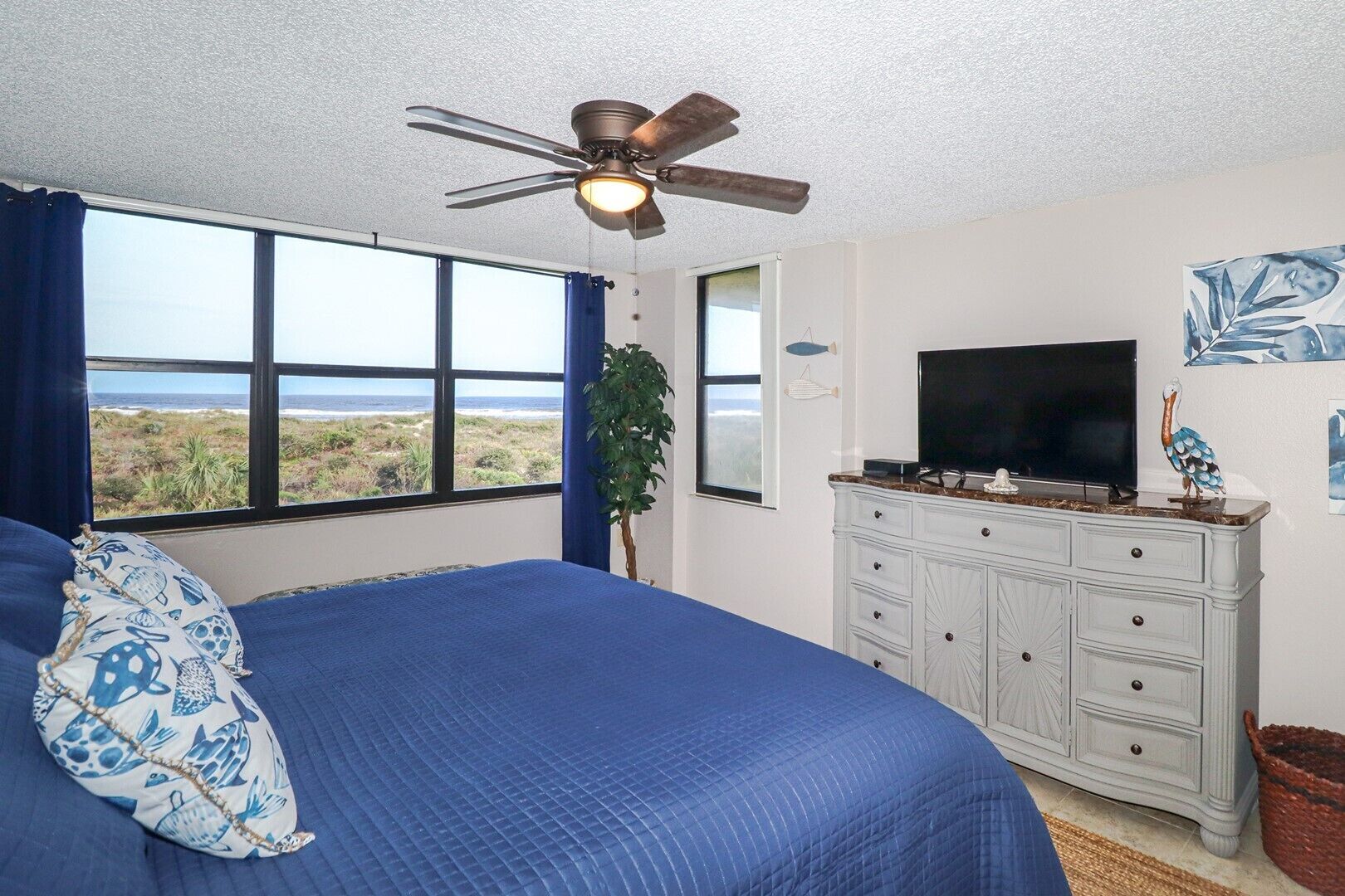 A bedroom with a large bed, a ceiling fan, a TV, and a view of the beach.