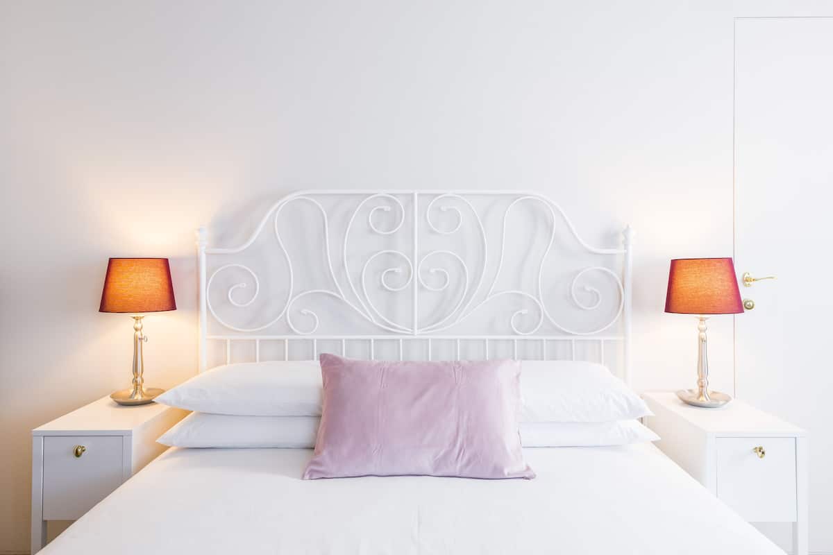 A neatly made bed with a white headboard, two bedside tables with lamps, and a single purple pillow.