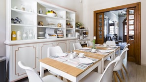 A dining area with a long table set for six, surrounded by white chairs. There are white cabinets with glass doors to the left, displaying various kitchen items and decorations. - Villa Savioli (Bologna)