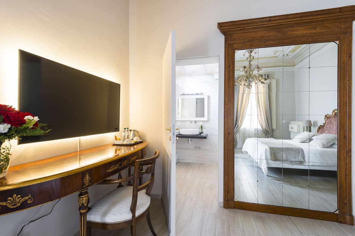 A hotel room with a large mirror, a bedside table with a vase of flowers, a bed with white bedding, and a bathroom visible through the mirror.