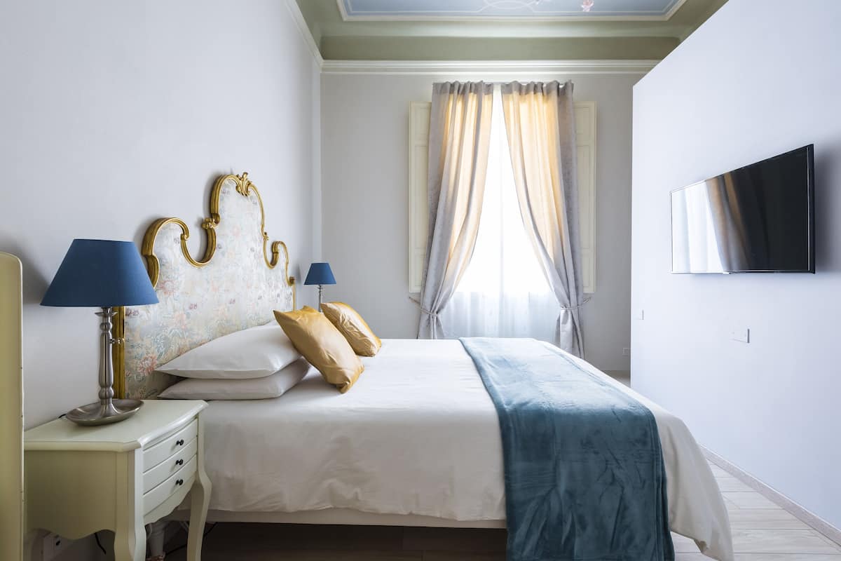 A neatly arranged bedroom with a bed, bedside lamps, a TV, and a window with curtains.