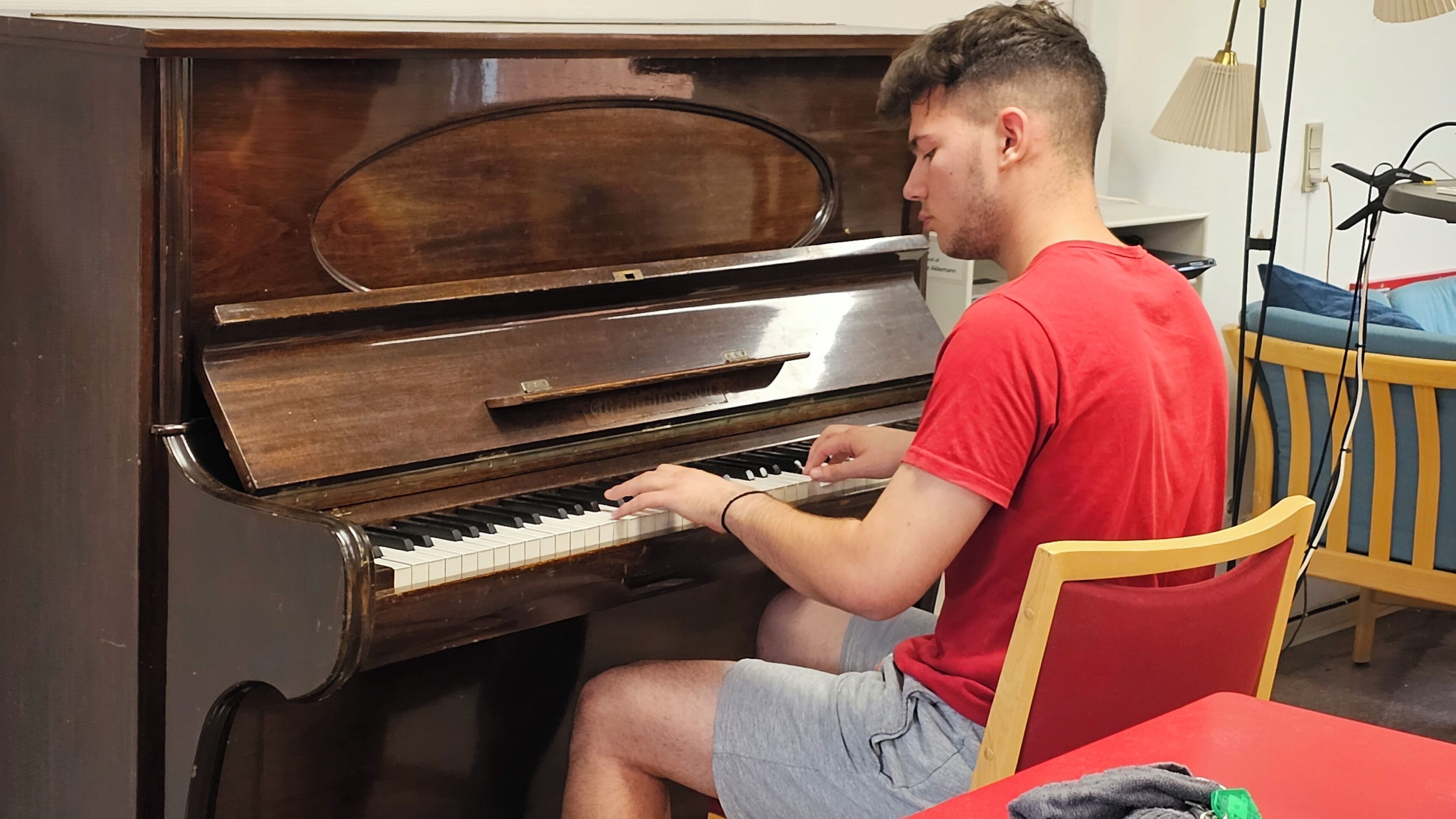 Uma pessoa sentada ao piano em um quarto com uma cadeira de madeira, uma mesa vermelha e um abajur ao fundo.