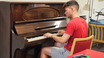 Uma pessoa sentada ao piano em um quarto com uma cadeira de madeira, uma mesa vermelha e um abajur ao fundo.