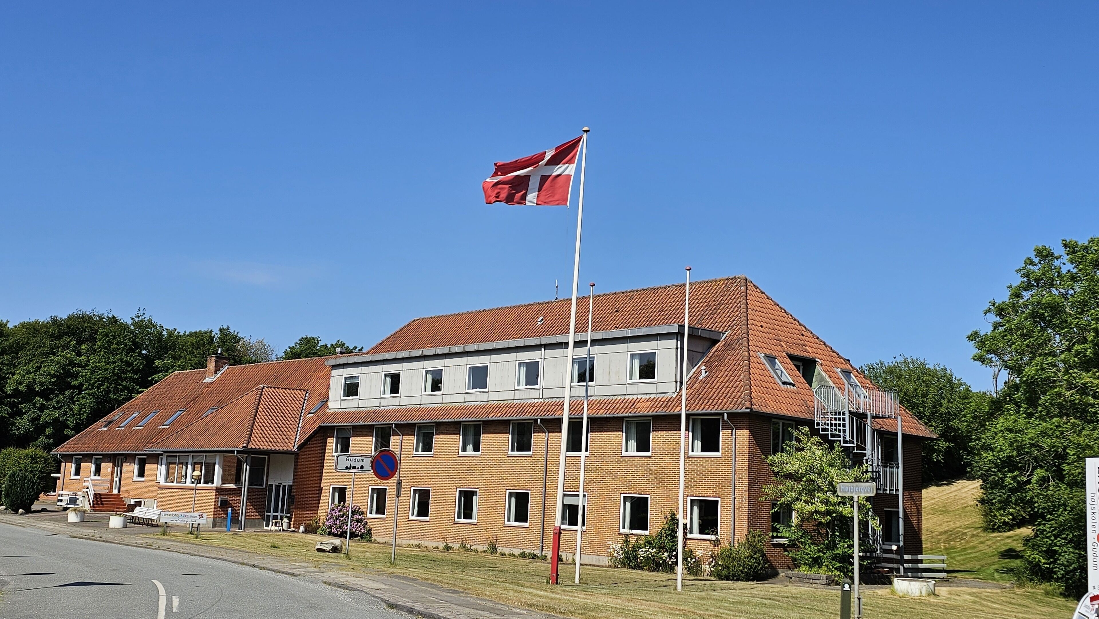 Un edificio de dos pisos con techo de tejas rojas, un asta con bandera y un cartel que indica 'Gudum' tanto en inglés como en otro idioma.