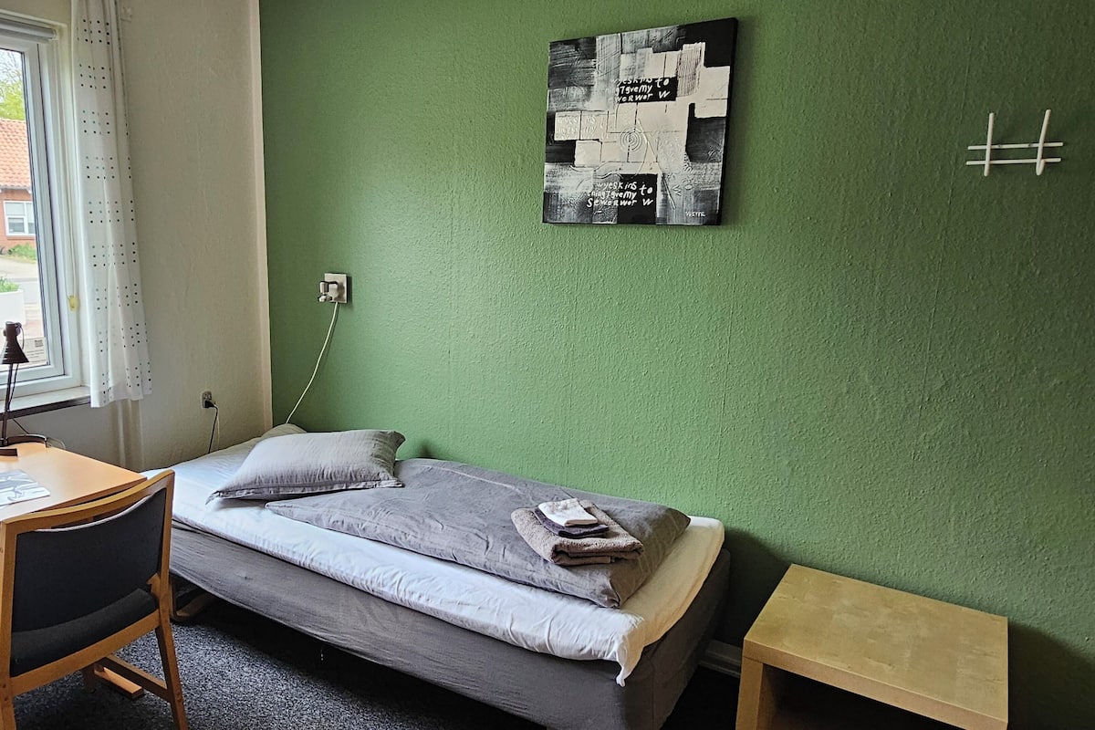 A small room with a bed, a desk, and a chair. There is a window with a view of buildings outside.