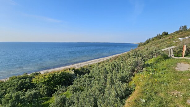 A coastal landscape with a beach, dense greenery, and a clear sky. - Gudum Park (Lemvig)