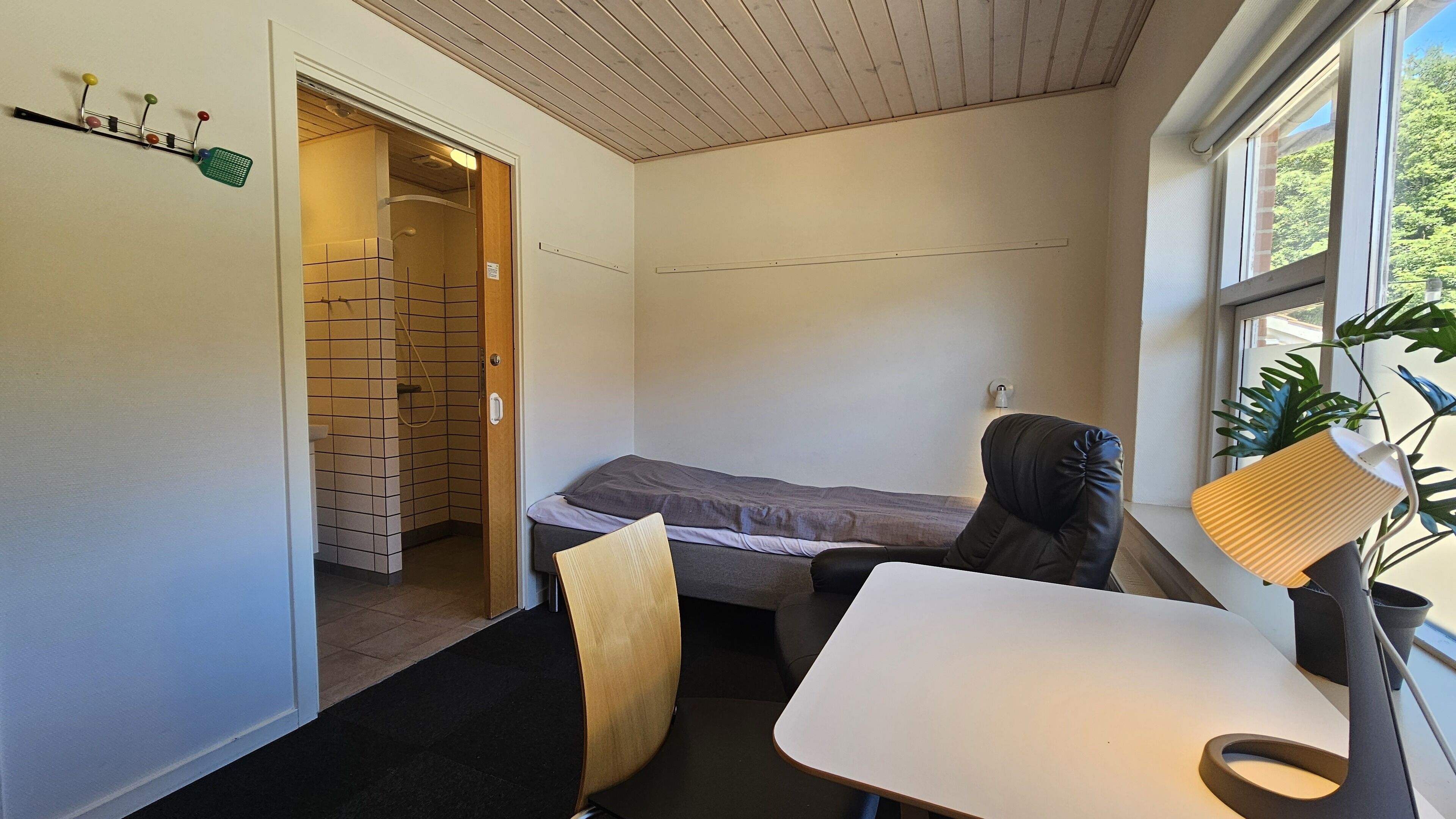 A compact room with a bed, a chair, a small table, a window, and a shower area.