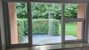 A view through a sliding glass door with a brick wall on one side and a garden with hedges and trees on the other. - Gudum Park (Lemvig)