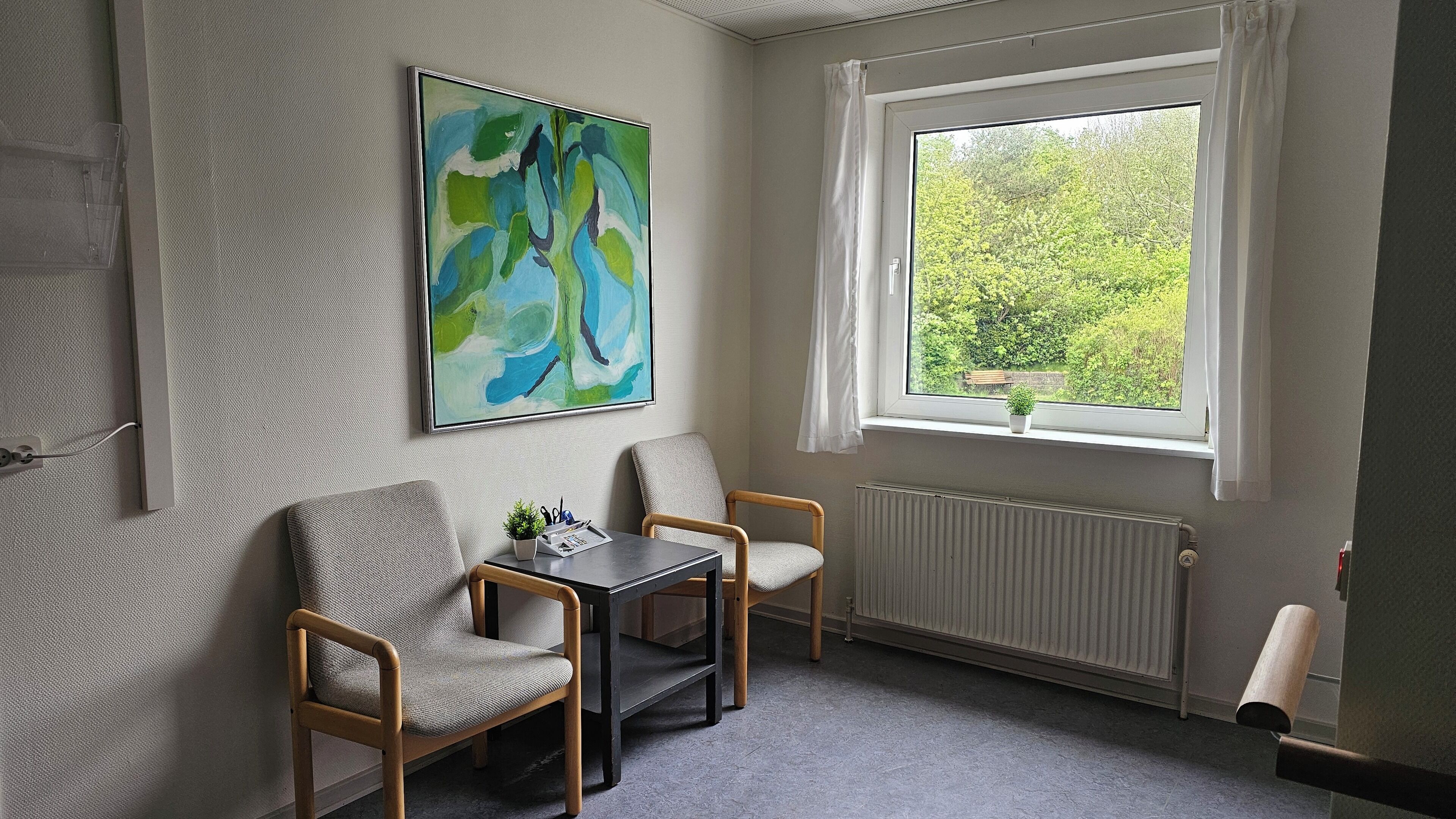 Et værelse med to stole, et bord, et billede, et vindue med udsigt til grøn natur og en radiator.