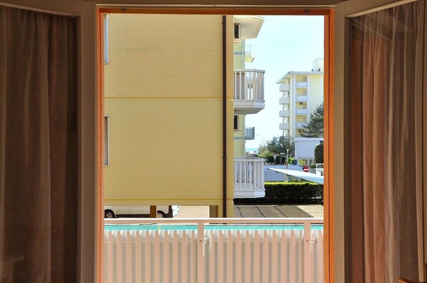 A view through an open door of a yellow building with a balcony, a white fence, and a street with buildings and a car in the background.