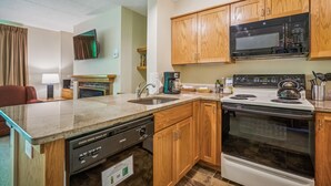 A compact kitchen with wooden cabinets, a microwave, a stove, and a dishwasher. - Pinnacle B04 Condo | Pool, Hot Tub | Golf Nearby (Killington)